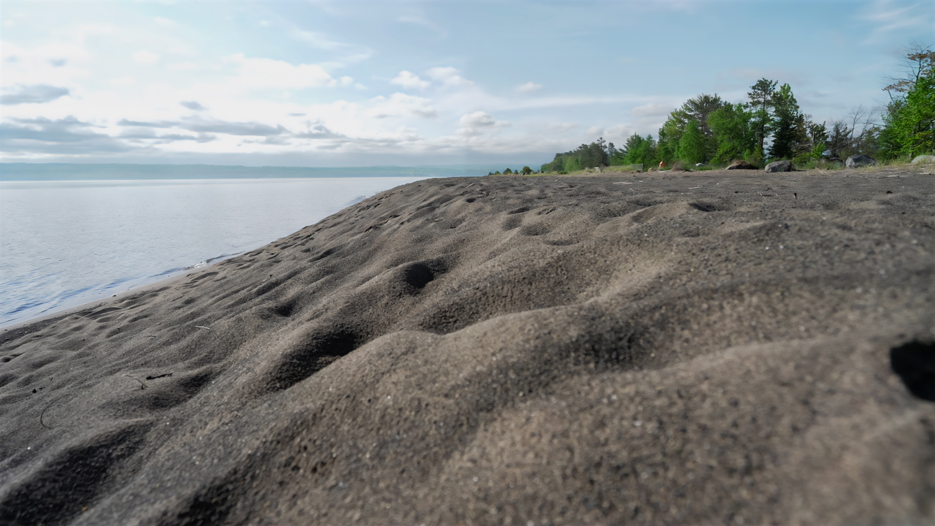 Close up of gray sand with water and trees in background.