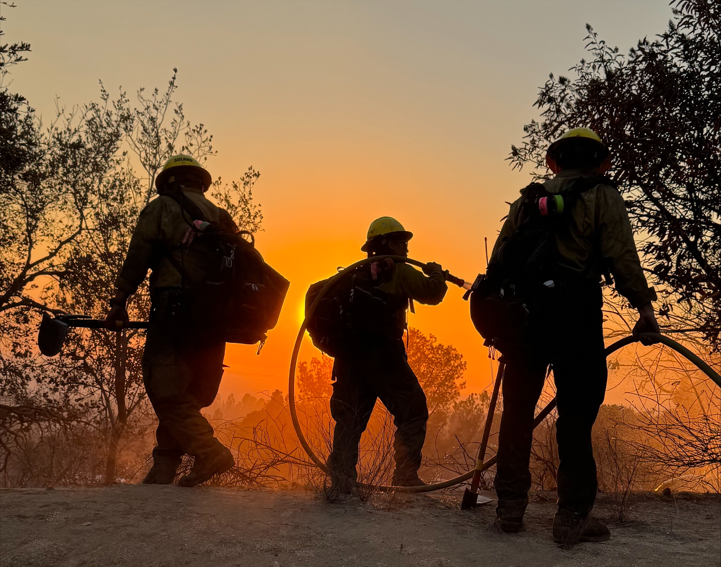 Image shows three wildland firefighters on a ridgeline with the sun setting behind them and the sky is orange with smoke from the Eaton wildfire.