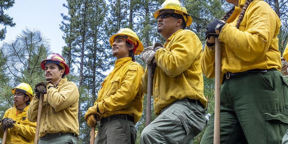 Inter-tribal youth standing in a half circle wearing wildland fire fighter safety closing and holding tools.