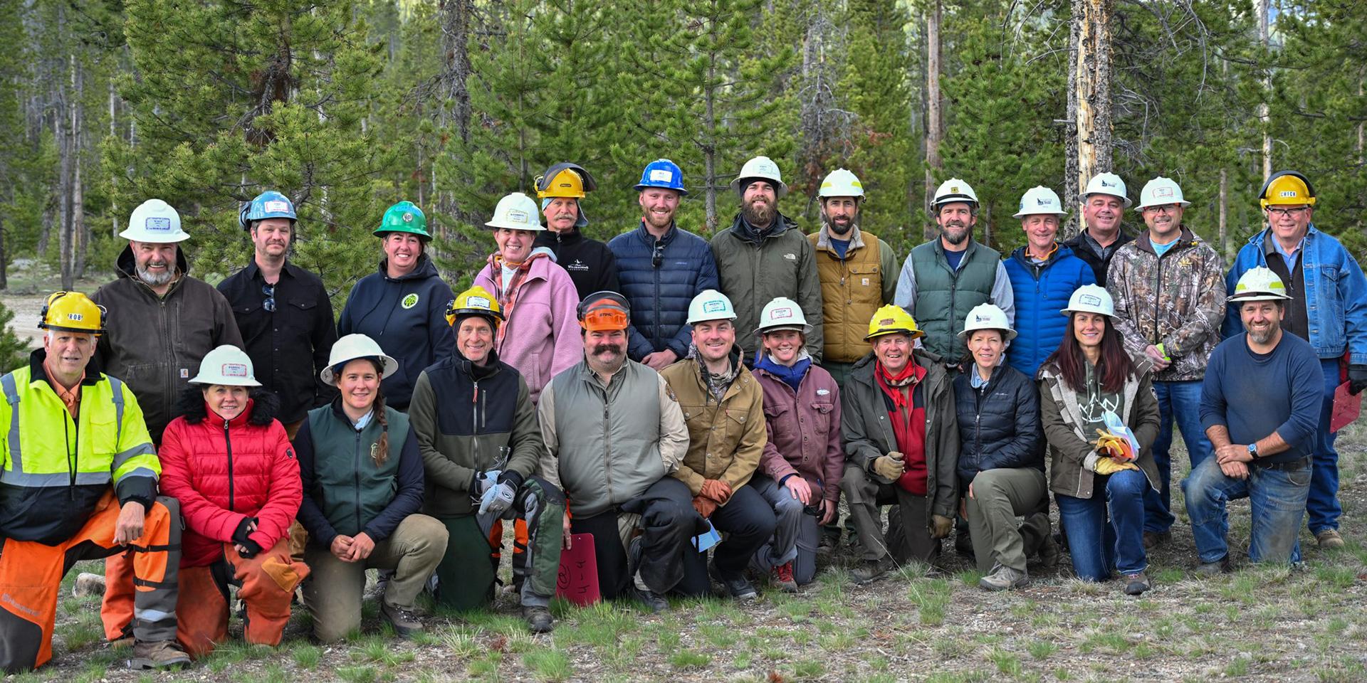 Twenty-four adults stand and kneel next to each other for a group photo in front of large green trees wearing hardhats and warm clothes.
