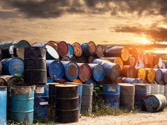 Rows of old 50 gallon metal chemical drums, some on their side while others are on end. A beautiful sunset shines in the distant sky.