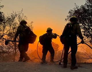 Image shows three wildland firefighters on a ridgeline with the sun setting behind them and the sky is orange with smoke from the Eaton wildfire.