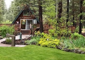 Old Station Visitor Center. A-frame wood building with wooden porch in front, surrounded by green grass, wildflowers, and tall pine trees. Pathway in center leads to porch and steps.