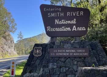 A wooden sign sits in on the side of the highway, welcoming travelers into the Smith River National Recreation Area. 